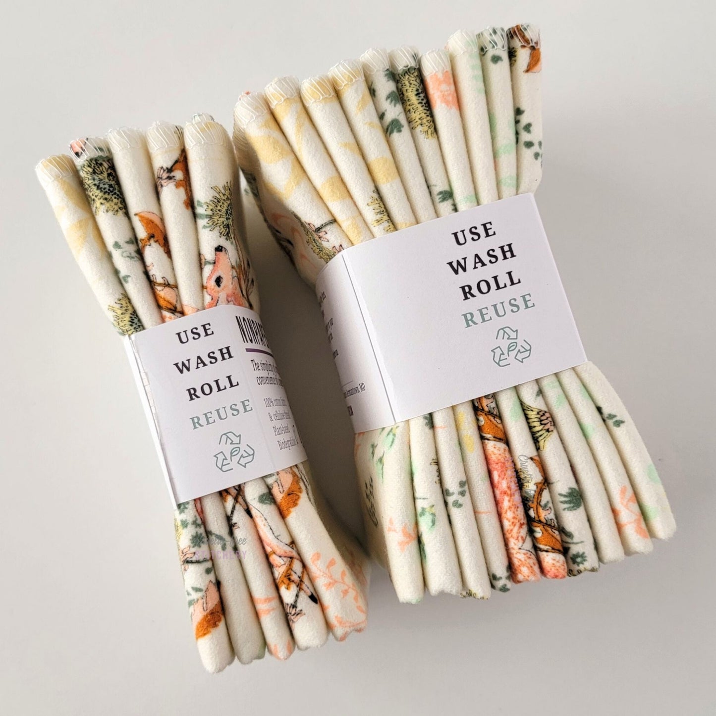 Two packages of the Dandelion Doe NonPaper Towels, shown on their side to display the 5-pack and 10-packs. The side of the paper tag says "use, wash, roll, reuse" and has the recycling logo with a leaf in the center, to symbolize they are biodegradable.