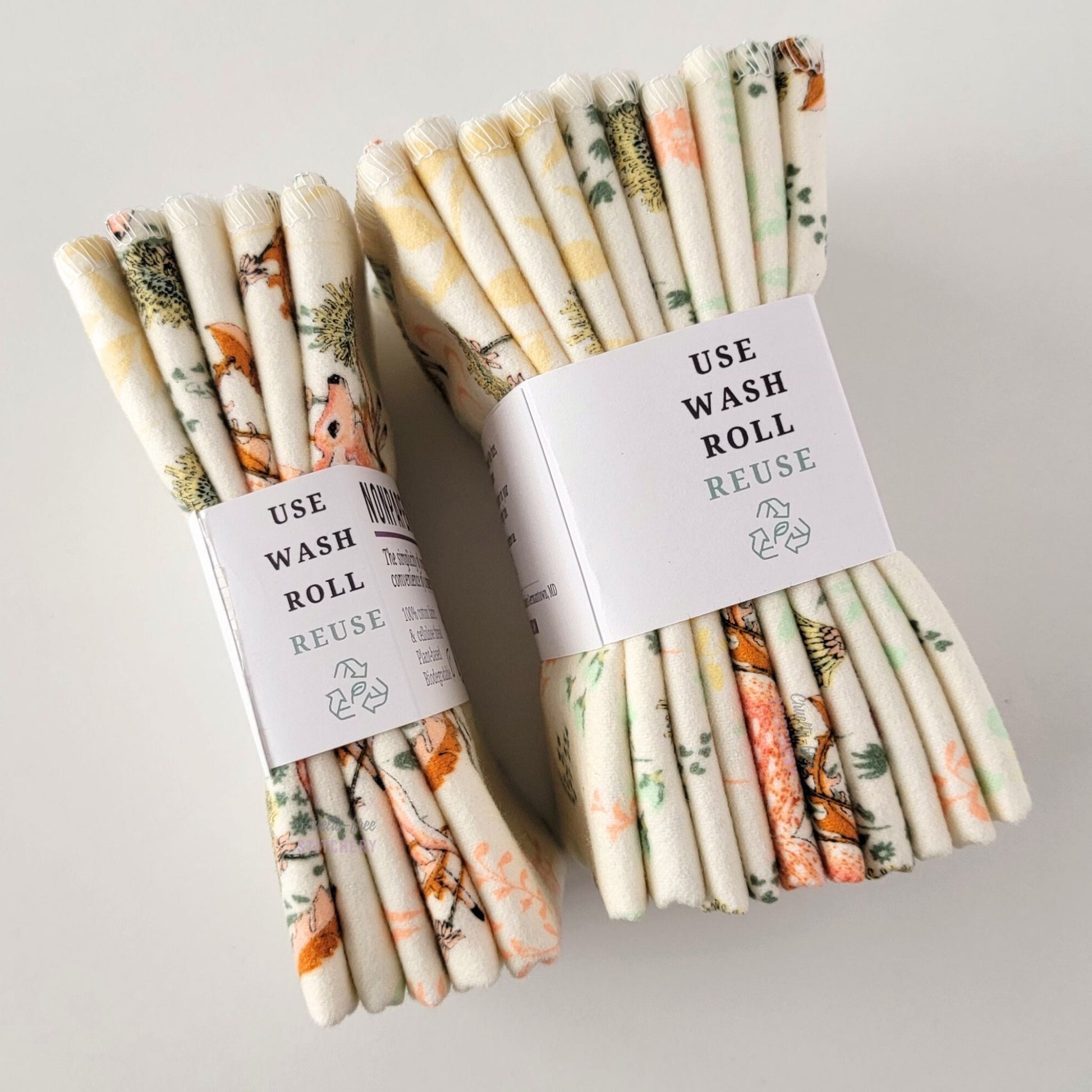 Two packages of the Dandelion Doe NonPaper Towels, shown on their side to display the 5-pack and 10-packs. The side of the paper tag says "use, wash, roll, reuse" and has the recycling logo with a leaf in the center, to symbolize they are biodegradable.