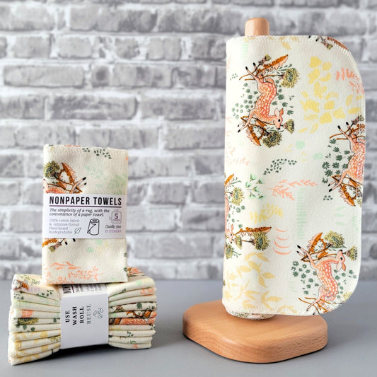 Dandelion Doe NonPaper Towels, displayed on a wooden paper towel holder next to packaged bundles. The print is a cream color background, with motifs of spotted deer in front of dandelions, with various pastel leaves and foliage around them. 