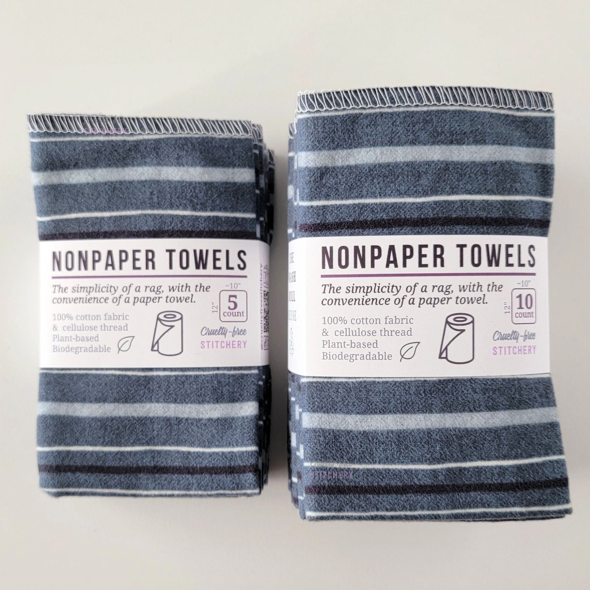 Two packages of the Deep Blue Stripe NonPaper Towels, one 5-pack and one 10-pack. Both wrapped with a paper tag, The tag also has the fiber content, a graphic of a paper towel roll, the Cruelty-Free Stitchery logo, and a rectangle with rounded corners that has the dimensions on the outside, 10x12", and the number of towels inside.