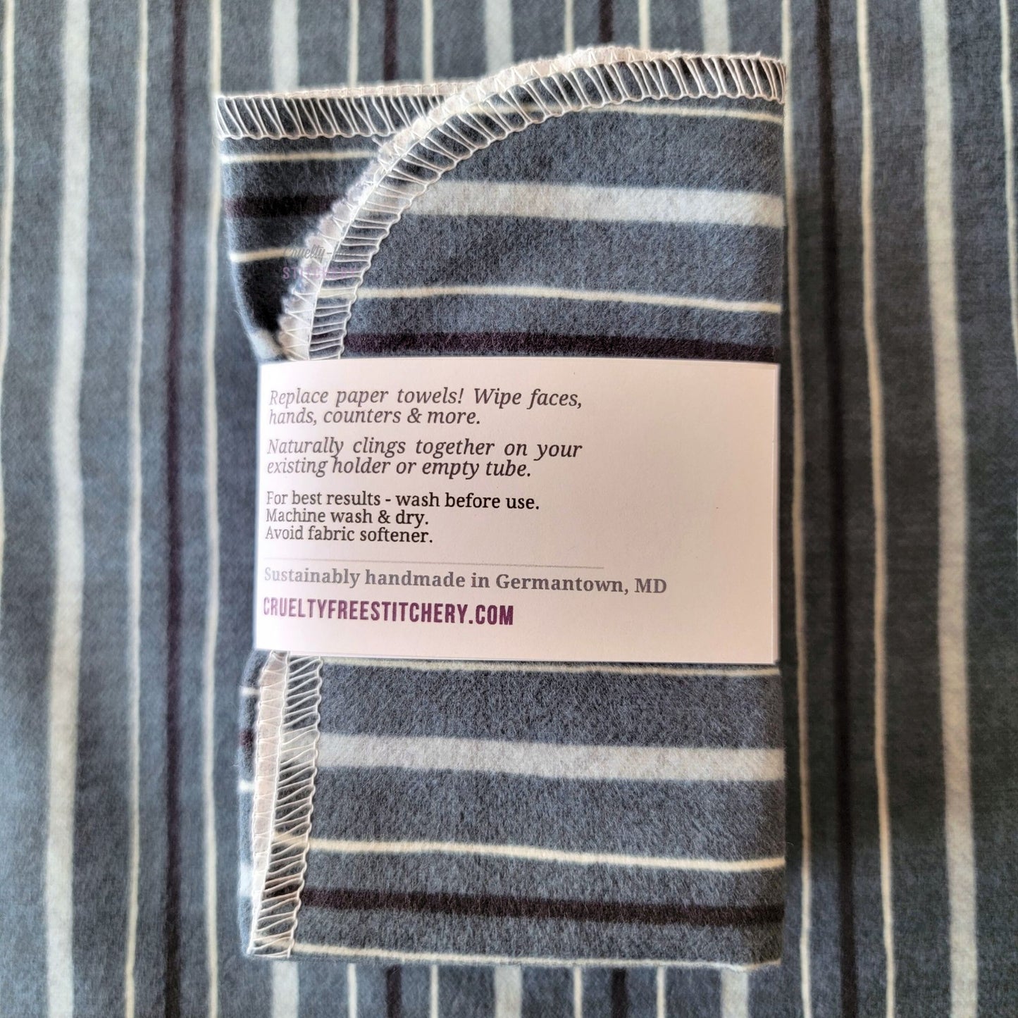 The back of a packaged bundle of the Deep Blue Stripe NonPaper Towels. The tag reads "Replace paper towels! Wipe faces, hands, counters, and more. Naturally clings together on your existing holder or empty tube. For best results - wash before use. Machine wash and dry. Avoid fabric softener. Sustainable handmade in Germantown, Maryland. CrueltyFreeStitchery dot com."