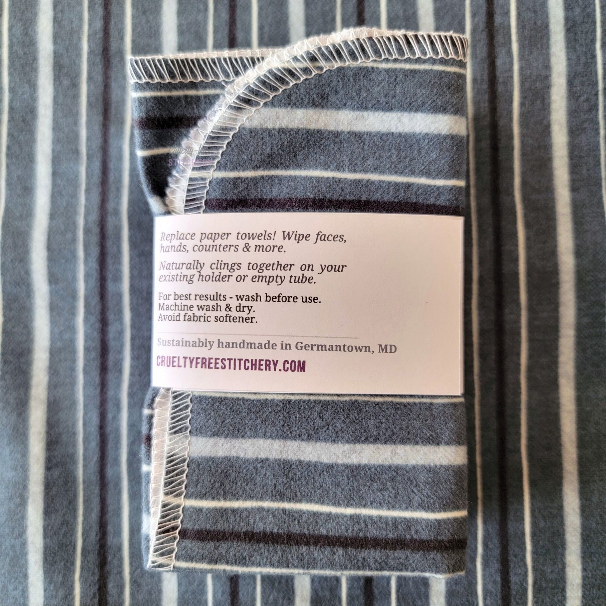 The back of a packaged bundle of the Deep Blue Stripe NonPaper Towels. The tag reads "Replace paper towels! Wipe faces, hands, counters, and more. Naturally clings together on your existing holder or empty tube. For best results - wash before use. Machine wash and dry. Avoid fabric softener. Sustainable handmade in Germantown, Maryland. CrueltyFreeStitchery dot com."