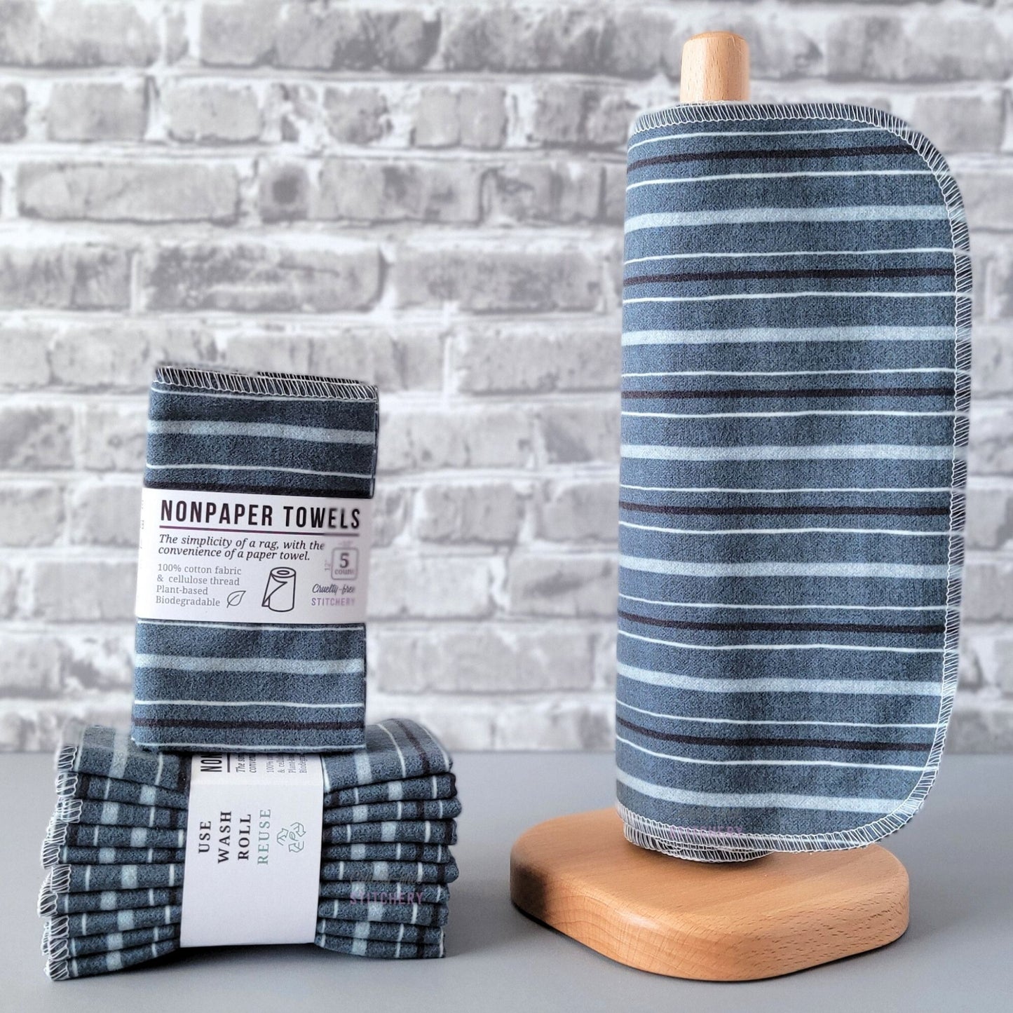 Deep Blue Stripe NonPaper Towels, displayed on a wooden paper towel holder next to packaged bundles. The print is an indigo denim color, with a pattern of 3 lighter blue stripes and one darker blue stripe. The stripe lines are going horizontally on the paper towel holder.