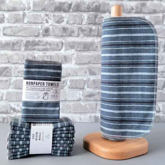 Deep Blue Stripe NonPaper Towels, displayed on a wooden paper towel holder next to packaged bundles. The print is an indigo denim color, with a pattern of 3 lighter blue stripes and one darker blue stripe. The stripe lines are going horizontally on the paper towel holder.
