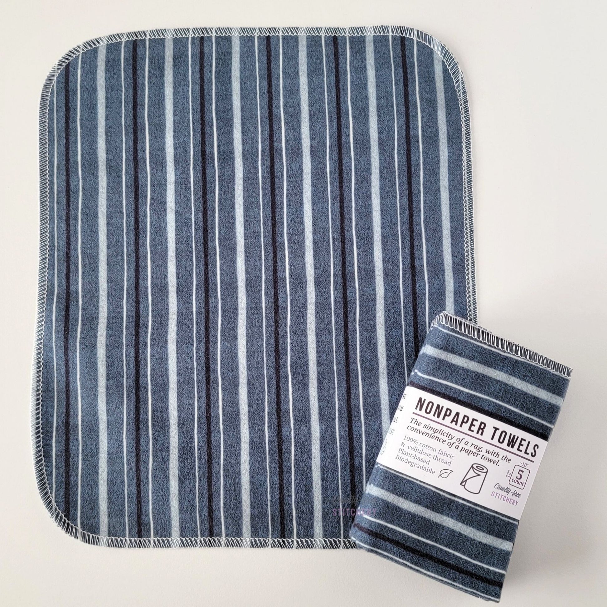 An unfolded Deep Blue Stripe NonPaper Towel, next to a bundled 5-pack. The towel is a rectangle with rounded corners, stitched around the edges with white thread. The bundled pack is folded up with a paper wrapper that says "NonPaper Towels, the simplicity of a rag with the convenience of a paper towel"
