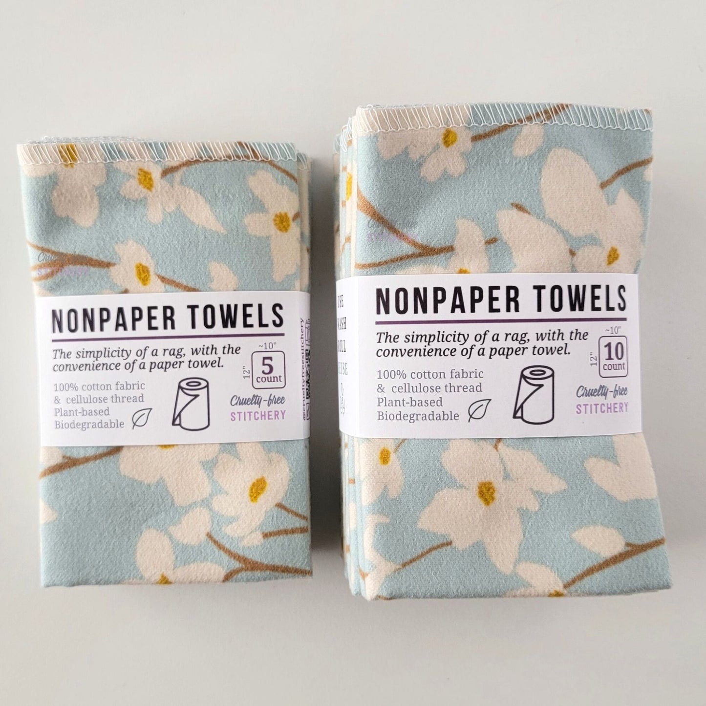 Two packages of the Dogwood Sky NonPaper Towels, one 5-pack and one 10-pack. Both wrapped with a paper tag, The tag also has the fiber content, a graphic of a paper towel roll, the Cruelty-Free Stitchery logo, and a rectangle with rounded corners that has the dimensions on the outside, 10x12", and the number of towels inside.