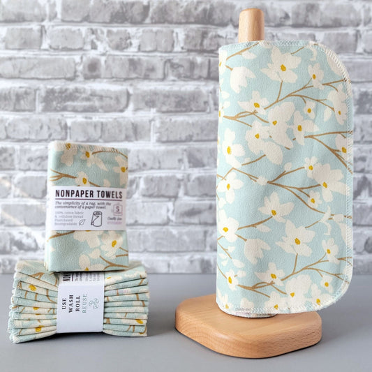 Dogwood Sky NonPaper Towels, displayed on a wooden paper towel holder next to packaged bundles. The print is a pale sky blue with off-white dogwood flowers growing on brown stems. 