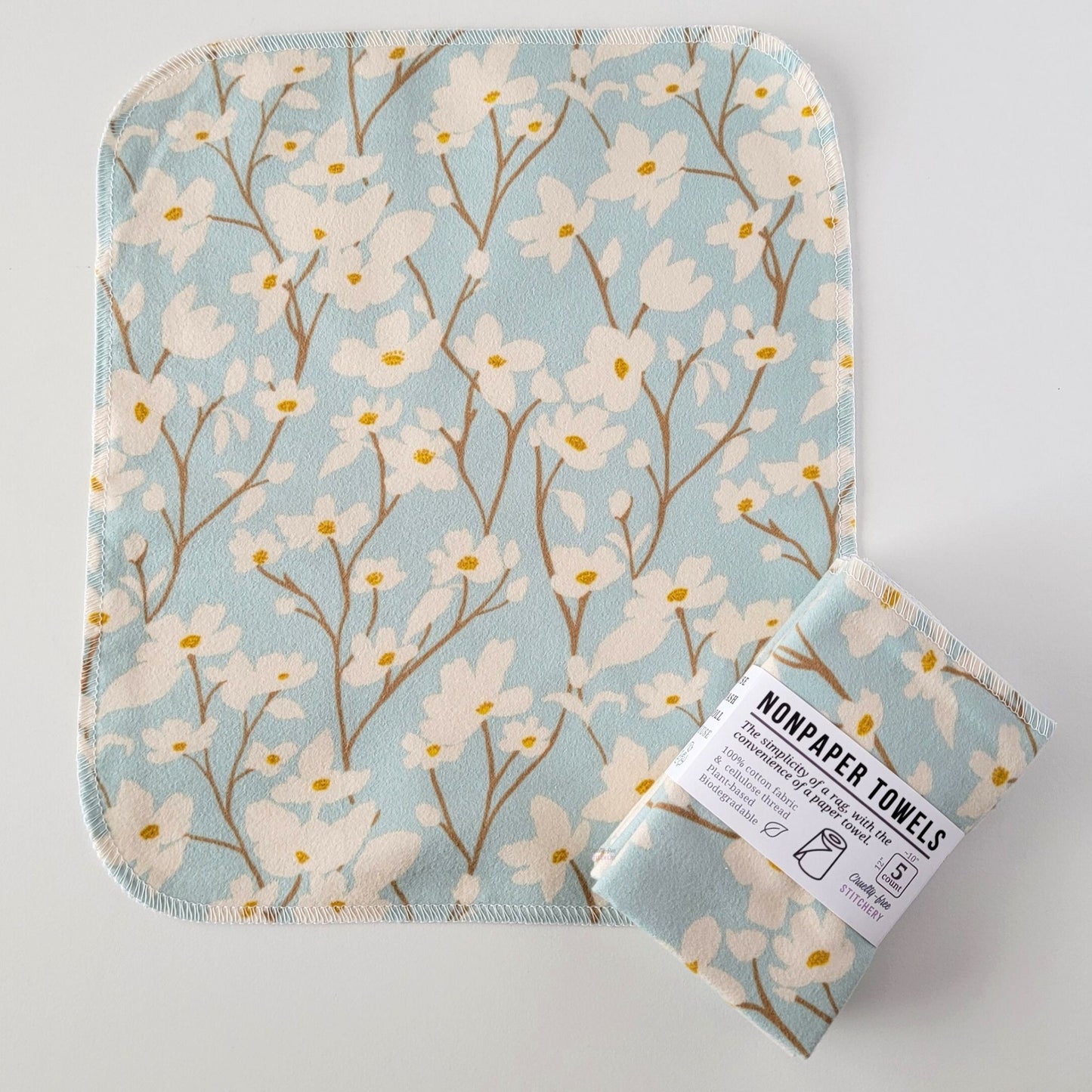 An unfolded Dogwood Sky NonPaper Towel, next to a bundled 5-pack. The towel is a rectangle with rounded corners, stitched around the edges with white thread. The bundled pack is folded up with a paper wrapper that says "NonPaper Towels, the simplicity of a rag with the convenience of a paper towel"