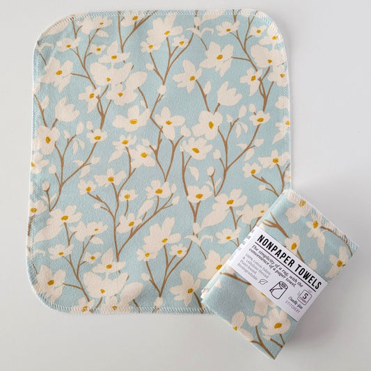 An unfolded Dogwood Sky NonPaper Towel, next to a bundled 5-pack. The towel is a rectangle with rounded corners, stitched around the edges with white thread. The bundled pack is folded up with a paper wrapper that says "NonPaper Towels, the simplicity of a rag with the convenience of a paper towel"