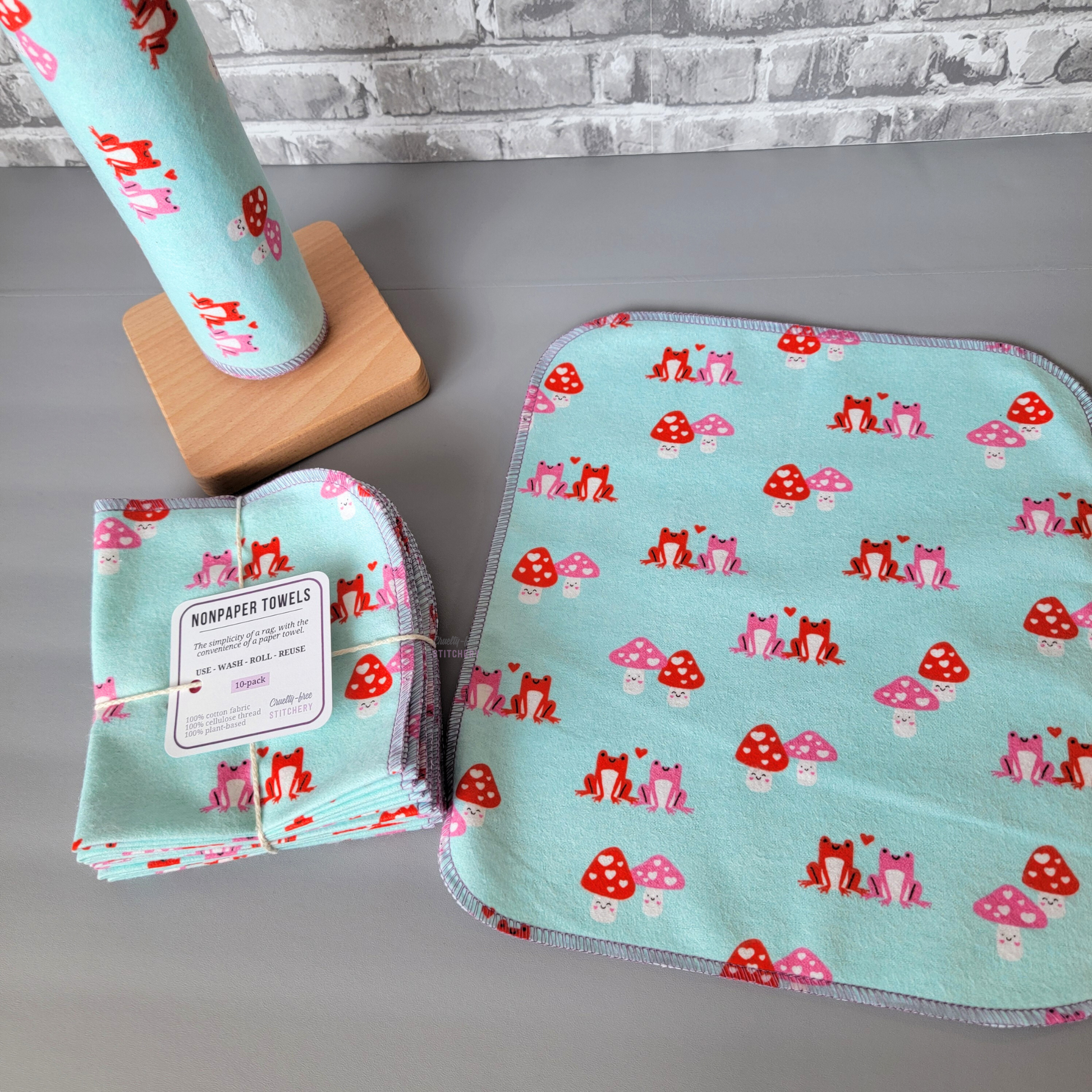 The blue frog mushroom print NonPaper Towels - one laid flat to show the print and size. The frog and mushroom couples are approximately 2 inches wide and evenly spaced. The edges of the towel are stitched with lilac purple thread with rounded corners. Next to it are a bundled pack and some on the wooden paper towel holder.