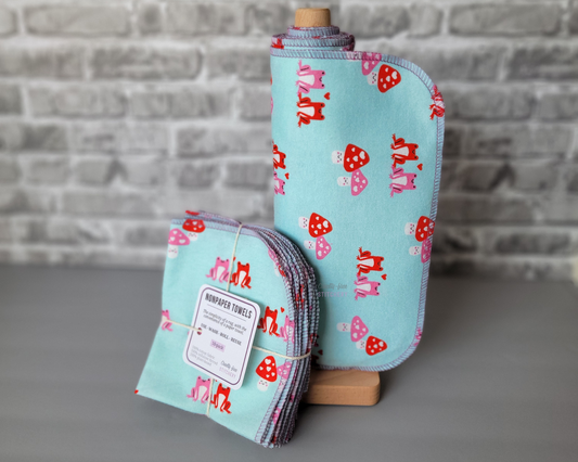 A set of NonPaper Towels - light aqua blue with small pink and red frogs and pink and red mushrooms with faces in a Valentine's day style. Some are rolled onto a wooden holder, next to a bundled pack tied with string and a small paper tag. 