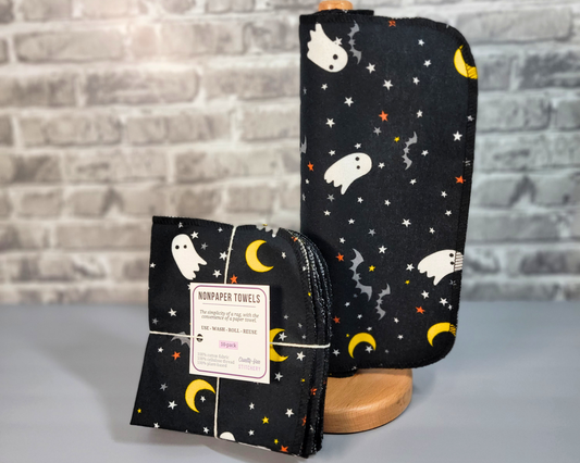 A roll of black with ghosts print NonPaper Towels next to a bundled pack. The towels have small white cartoonish ghosts, gray bats, moons, and tiny stars. Stitched in black thread and rounded corners.