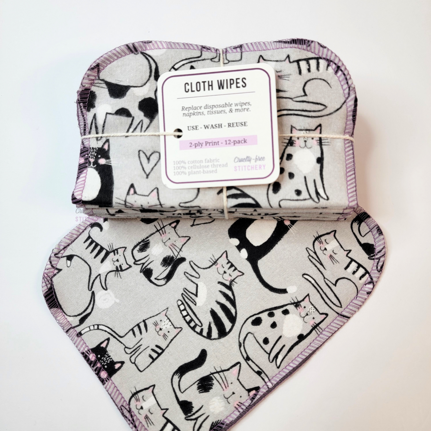 Gray cats print cloth wipes, a bundled pack that is folded in half and tied with string and a small paper tag, on top of a wipe laid flat. The tag reads "cloth wipes. Replace disposable wipes, napkins, tissues, and more. Use, wash, reuse. 2-ply print, 12-pack. 100% cotton fabric, 100% cellulose thread. 100% plant based. Cruelty-Free Stitchery"