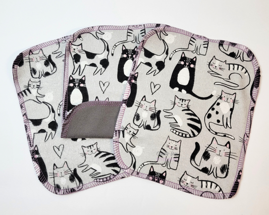 Gray Cats Cloth Wipes. Three wipes arranged with one in the middle folded up to show the back is a darker gray. The front is light gray with sketched black and white cats in various positions. Edges stitched in a lilac purple with rounded corners.