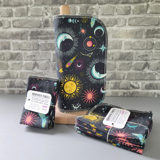 Gray celestial NonPaper Towels, displayed on a wooden paper towel holder, with bundled 5 and 10-packs sitting next to it. The towels have various motifs like suns, moons, stars, comets, saturn, moths, etc in shades of light aqua blue, yellow, and pink.