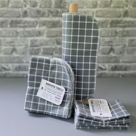 Gray Small Windowpane print NonPaper Towels. The print is a medium gray with white plaid squares about 3/4" wide. In the center, they are rolled onto a wooden paper towel holder, and there are bundled packs of 10 and 5 sitting next to the holder. The bundles are tied with string and have a small paper tag. The background is a gray brick wall and solid gray tabletop. 