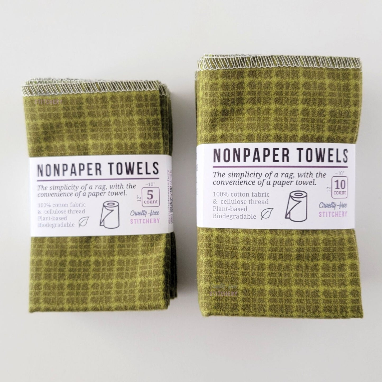 Two packages of the Green Kitchen Plaid NonPaper Towels, one 5-pack and one 10-pack. Both wrapped with a paper tag, The tag also has the fiber content, a graphic of a paper towel roll, the Cruelty-Free Stitchery logo, and a rectangle with rounded corners that has the dimensions on the outside, 10x12", and the number of towels inside.