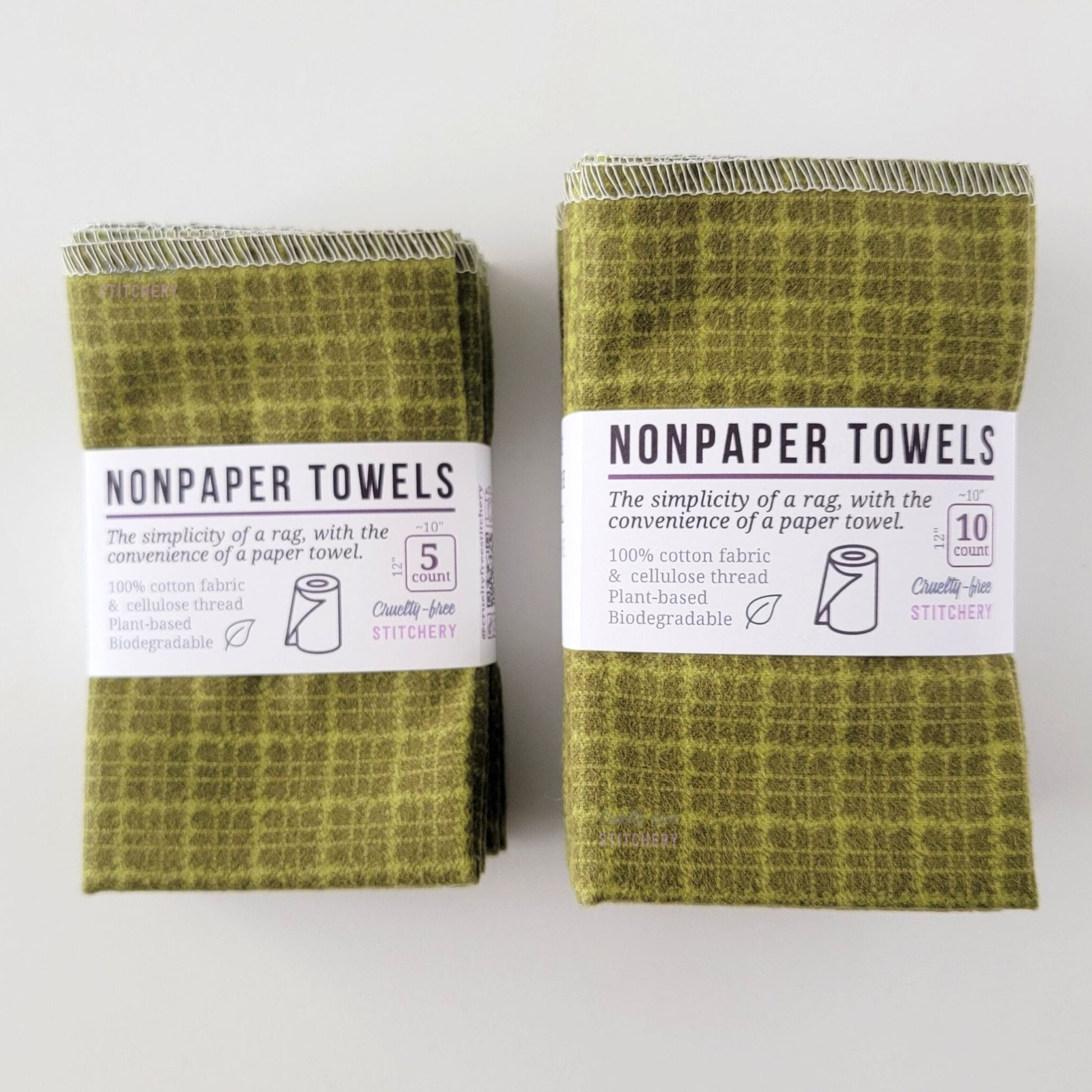 Two packages of the Green Kitchen Plaid NonPaper Towels, one 5-pack and one 10-pack. Both wrapped with a paper tag, The tag also has the fiber content, a graphic of a paper towel roll, the Cruelty-Free Stitchery logo, and a rectangle with rounded corners that has the dimensions on the outside, 10x12", and the number of towels inside.