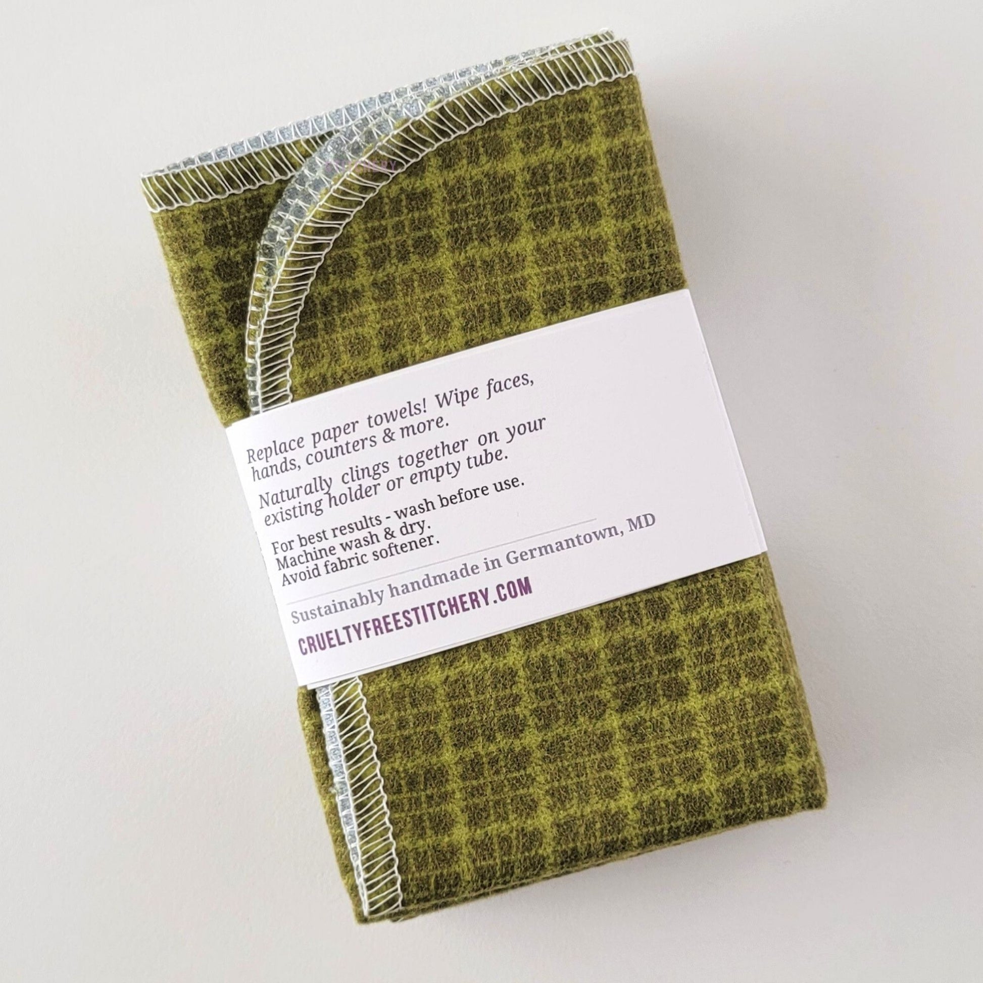 The back of a packaged bundle of the Green Kitchen Plaid NonPaper Towels. The tag reads "Replace paper towels! Wipe faces, hands, counters, and more. Naturally clings together on your existing holder or empty tube. For best results - wash before use. Machine wash and dry. Avoid fabric softener. Sustainable handmade in Germantown, Maryland. CrueltyFreeStitchery dot com."