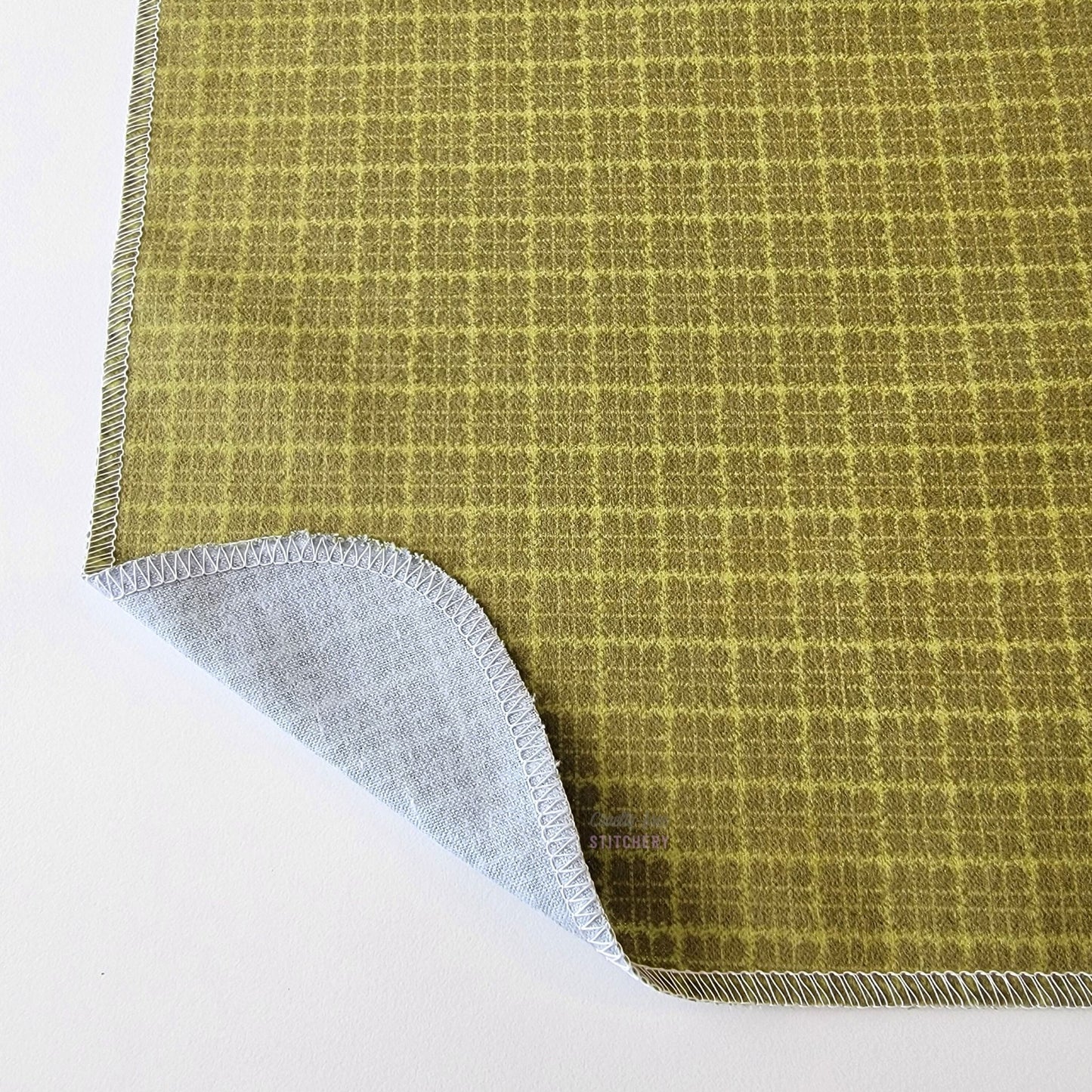 The Green Kitchen Plaid NonPaper Towel, shown close-up with the bottom corner folded back to show they are unprinted on the back side, and are one layer.