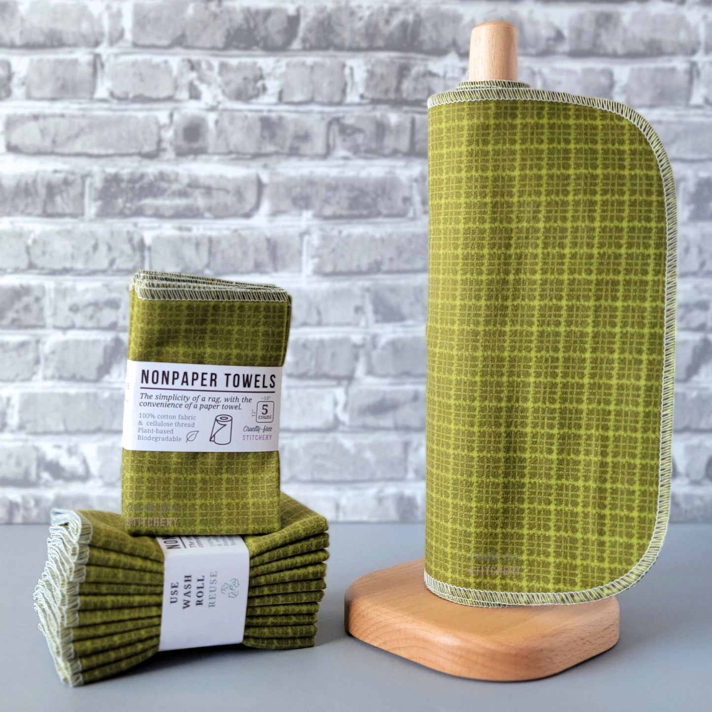 Green Kitchen Plaid NonPaper Towels, displayed on a wooden paper towel holder next to packaged bundles. They are an olive green with lighter green plaid lines. The lines are more scribbly when you look closer. The plaid squares are approximately half an inch.