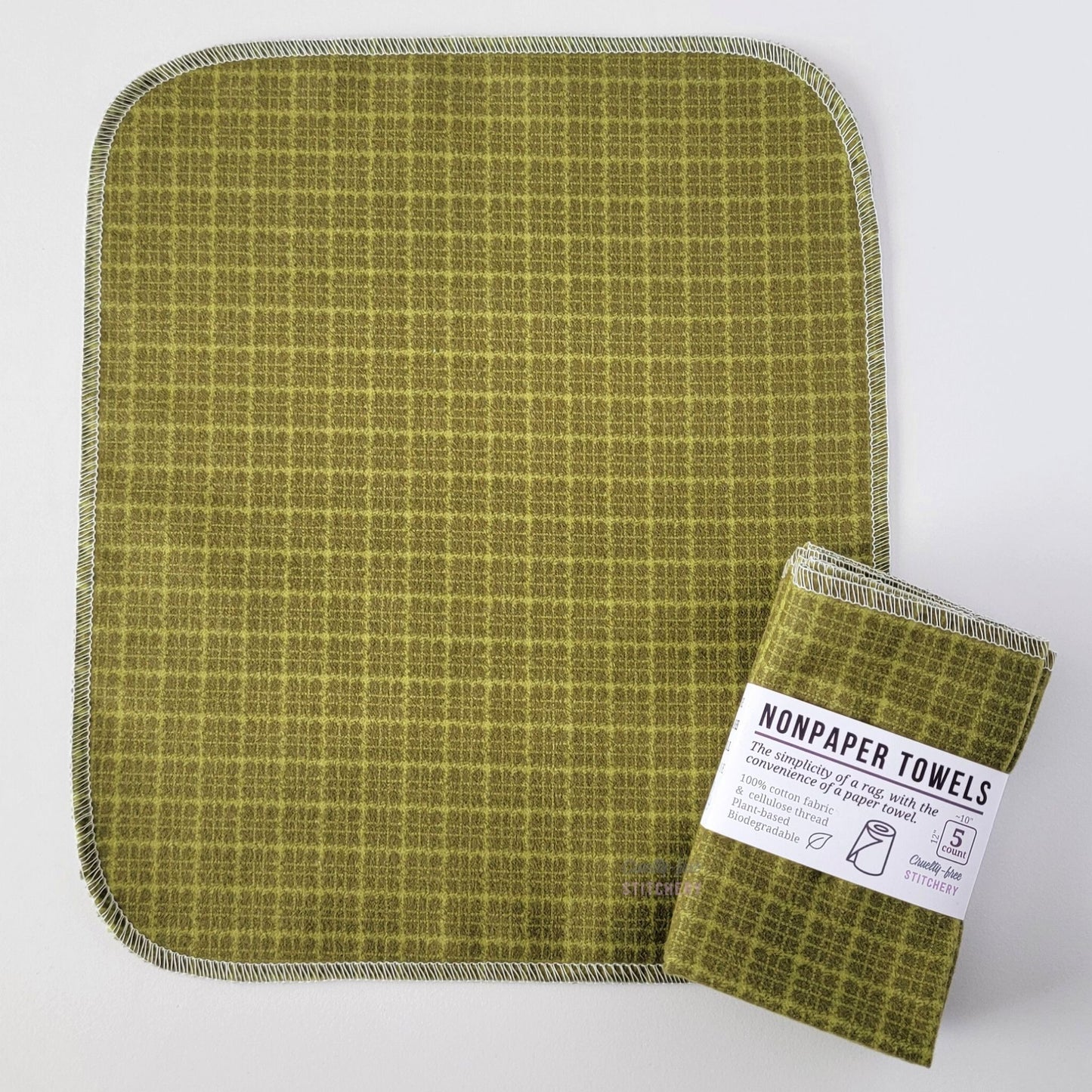 An unfolded Green Kitchen Plaid NonPaper Towel, next to a bundled 5-pack. The towel is a rectangle with rounded corners, stitched around the edges with white thread. The bundled pack is folded up with a paper wrapper that says "NonPaper Towels, the simplicity of a rag with the convenience of a paper towel"