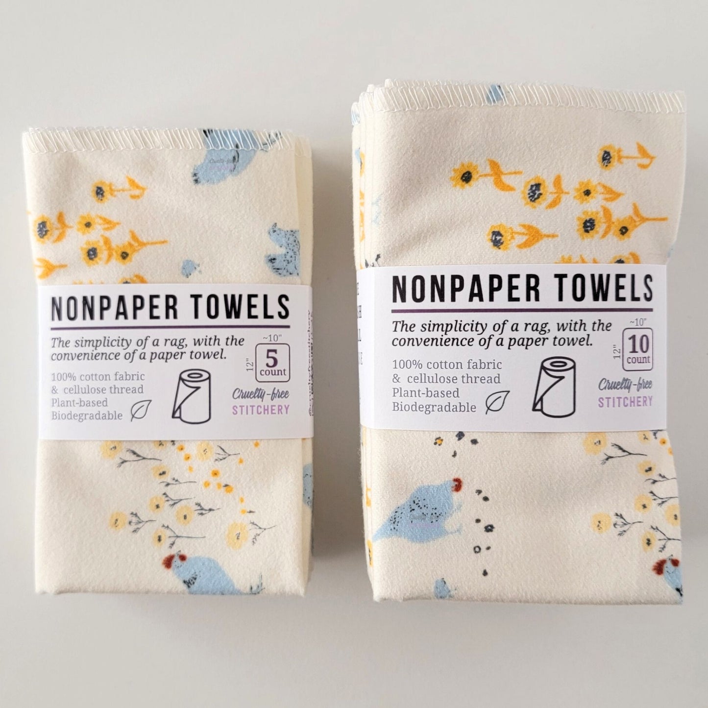 Two packages of the Hens and Sunflowers NonPaper Towels, one 5-pack and one 10-pack. Both wrapped with a paper tag, The tag also has the fiber content, a graphic of a paper towel roll, the Cruelty-Free Stitchery logo, and a rectangle with rounded corners that has the dimensions on the outside, 10x12", and the number of towels inside.