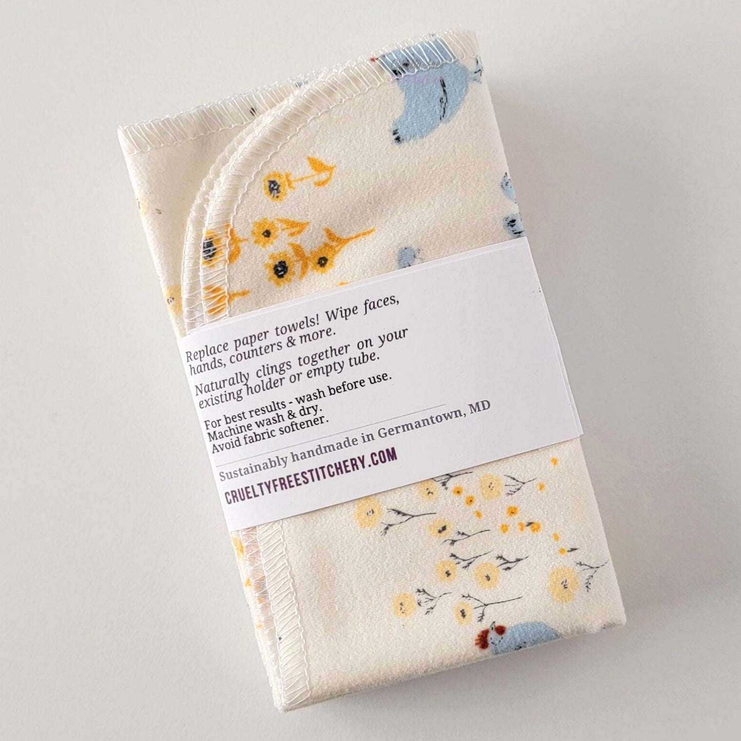 The back of a packaged bundle of the Hens and Sunflowers NonPaper Towels. The tag reads "Replace paper towels! Wipe faces, hands, counters, and more. Naturally clings together on your existing holder or empty tube. For best results - wash before use. Machine wash and dry. Avoid fabric softener. Sustainable handmade in Germantown, Maryland. CrueltyFreeStitchery dot com."