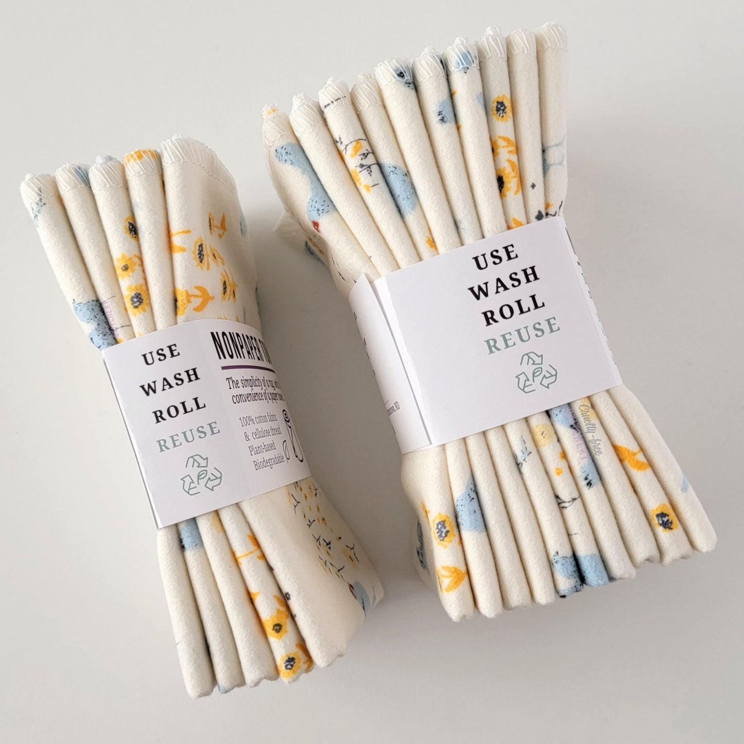 Two packages of the Hens and Sunflowers NonPaper Towels, shown on their side to display the 5-pack and 10-packs. The side of the paper tag says "use, wash, roll, reuse" and has the recycling logo with a leaf in the center, to symbolize they are biodegradable.