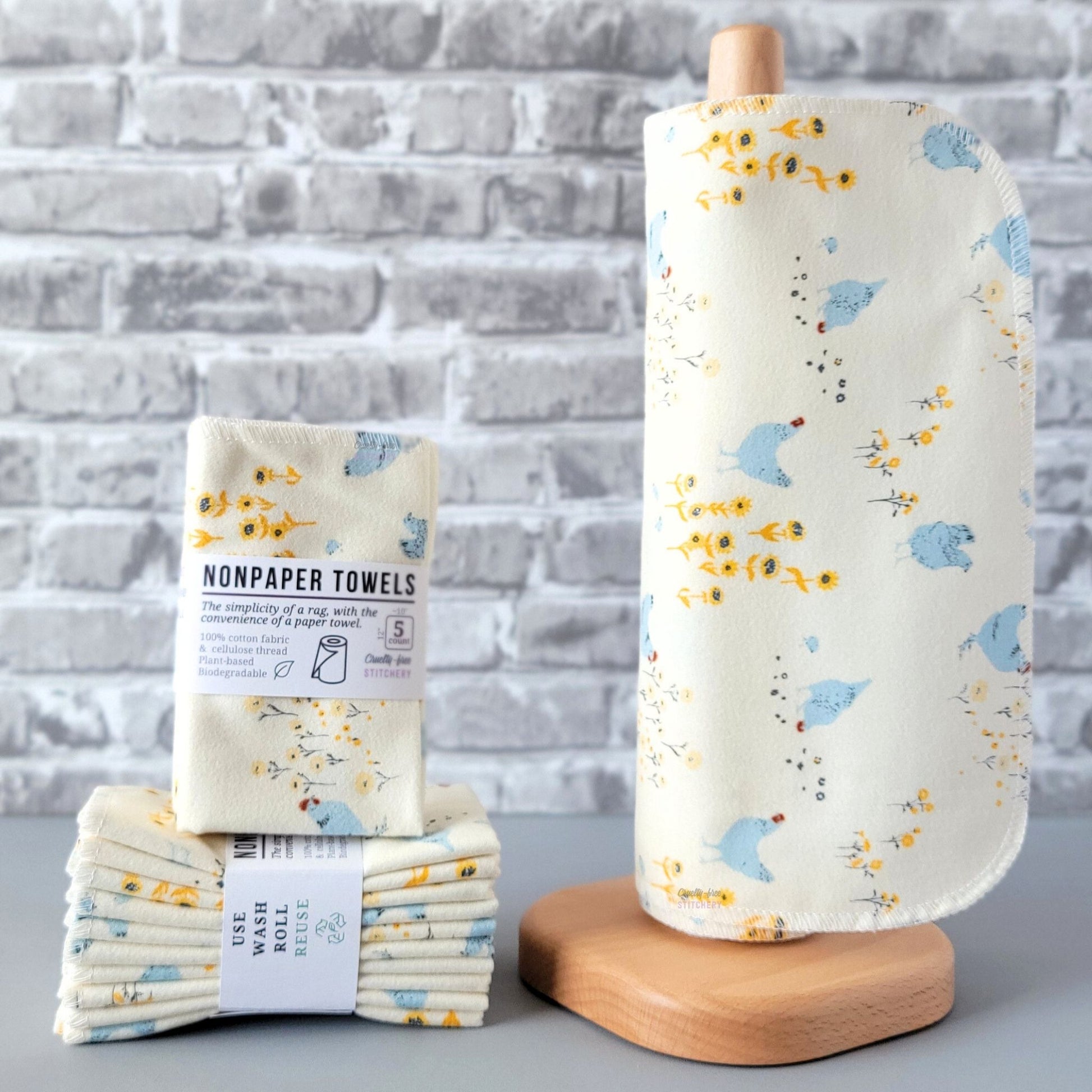 Hens and Sunflowers NonPaper Towels, displayed on a wooden paper towel holder next to packaged bundles. The print is an off-white or cream color, with light blue chickens scattered around. There are yellow sunflowers and tiny little blue chicks as well. The chickens are less than an inch tall, some are standing, some are pecking the ground. 