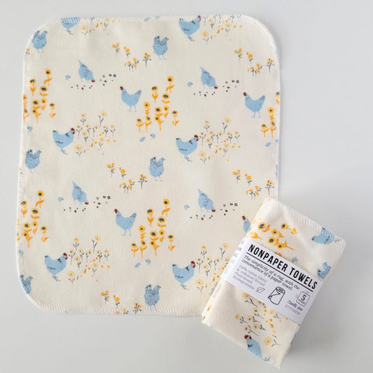 An unfolded Hens and Sunflowers NonPaper Towel, next to a bundled 5-pack. The towel is a rectangle with rounded corners, stitched around the edges with white thread. The bundled pack is folded up with a paper wrapper that says "NonPaper Towels, the simplicity of a rag with the convenience of a paper towel"