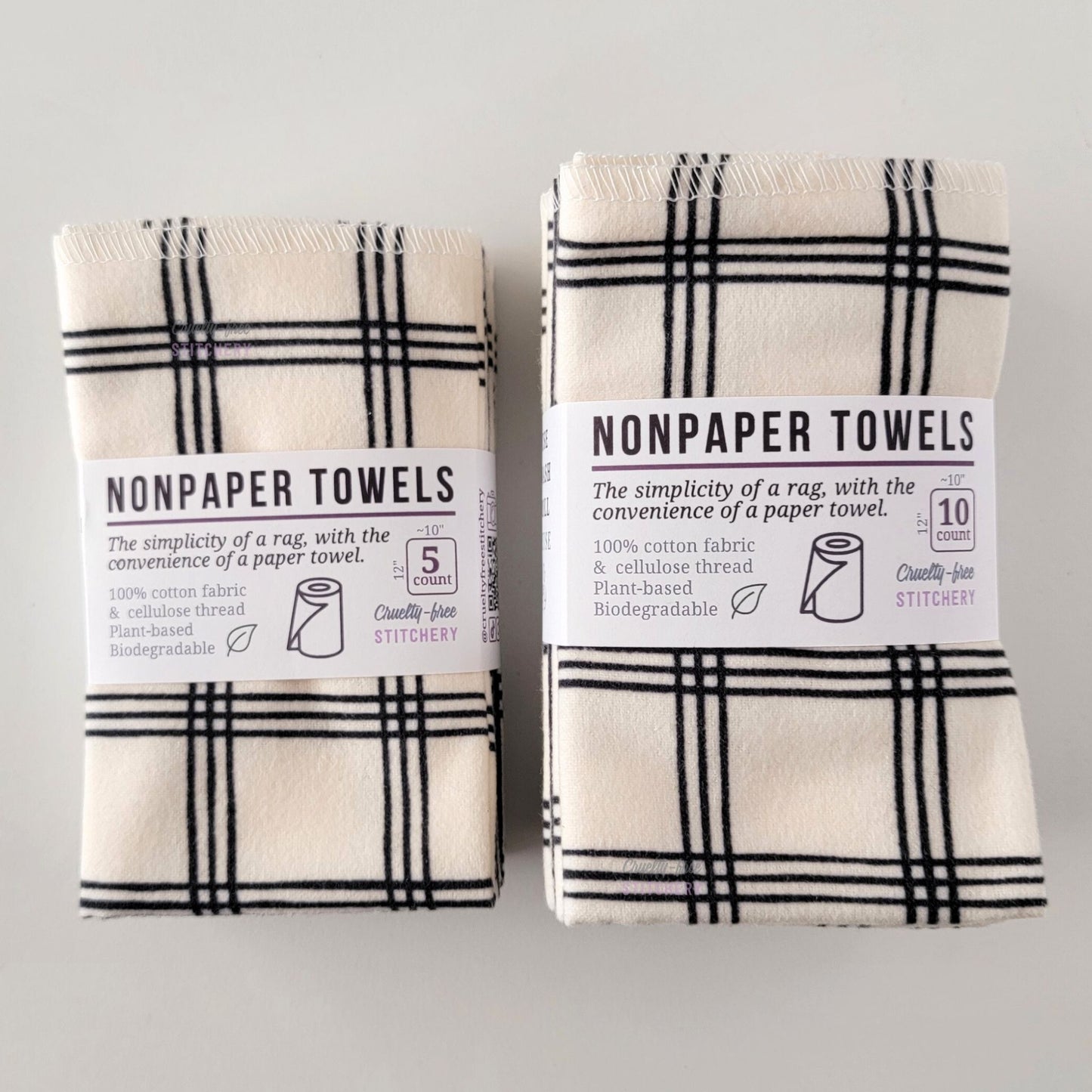 Two packages of the Ivory Black Crossroad NonPaper Towels, one 5-pack and one 10-pack. Both wrapped with a paper tag, The tag also has the fiber content, a graphic of a paper towel roll, the Cruelty-Free Stitchery logo, and a rectangle with rounded corners that has the dimensions on the outside, 10x12", and the number of towels inside.
