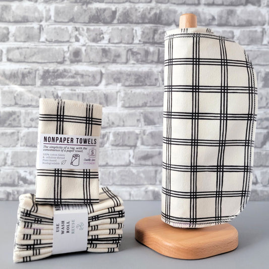Ivory Black Crossroad NonPaper Towels, displayed on a wooden paper towel holder next to packaged bundles. The print is a light ivory color, with a simple square plaid pattern that is made up of groups of three thin, black lines going vertically and horizontally. The square spaces between the grid lines are approximately an inch.