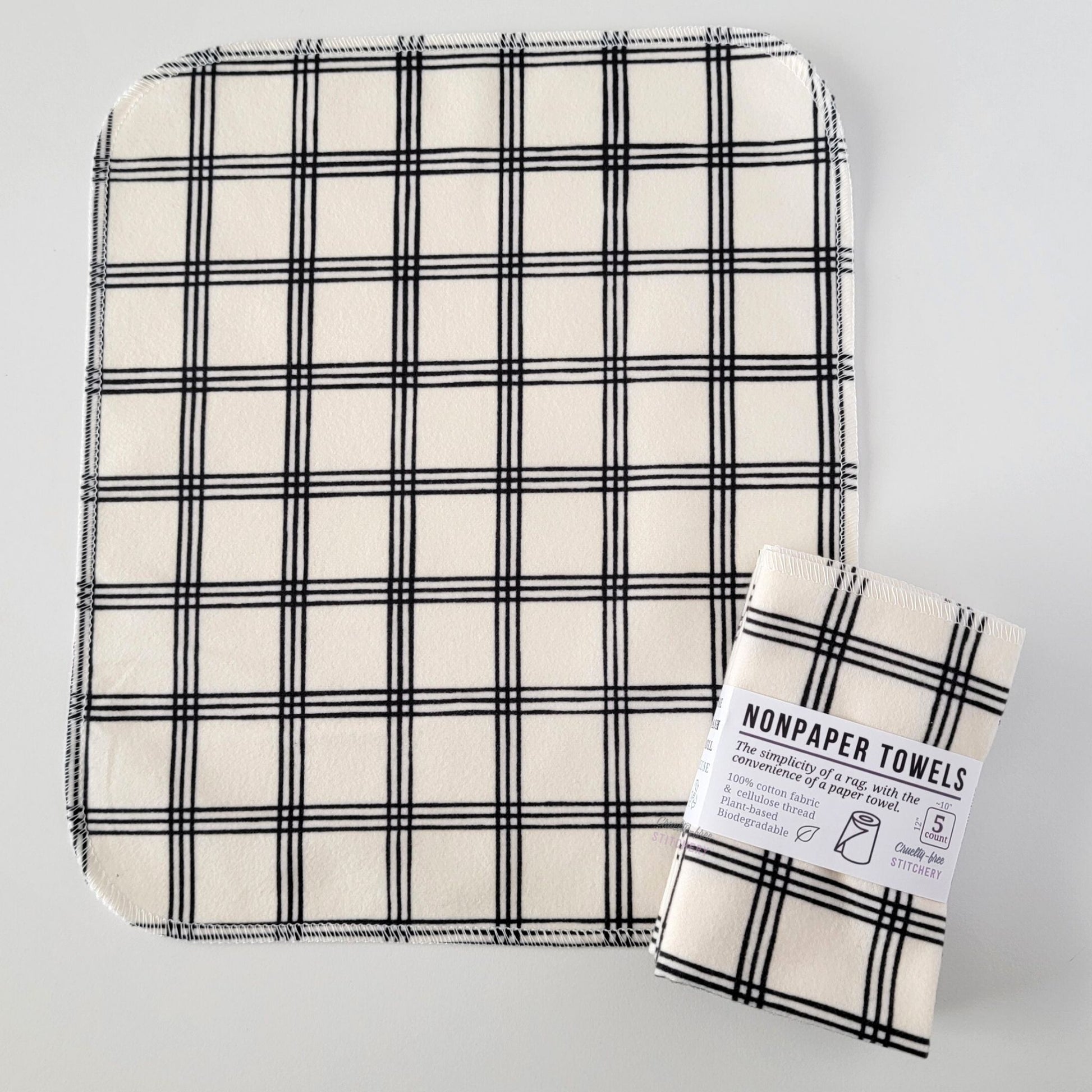 An unfolded Ivory Black Crossroad NonPaper Towel, next to a bundled 5-pack. The towel is a rectangle with rounded corners, stitched around the edges with white thread. The bundled pack is folded up with a paper wrapper that says "NonPaper Towels, the simplicity of a rag with the convenience of a paper towel"