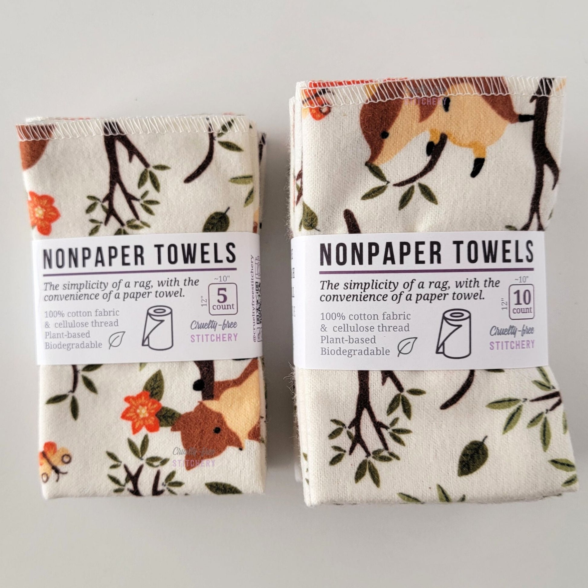 Two packages of the Ivory Foxes NonPaper Towels, one 5-pack and one 10-pack. Both wrapped with a paper tag, The tag also has the fiber content, a graphic of a paper towel roll, the Cruelty-Free Stitchery logo, and a rectangle with rounded corners that has the dimensions on the outside, 10x12", and the number of towels inside.