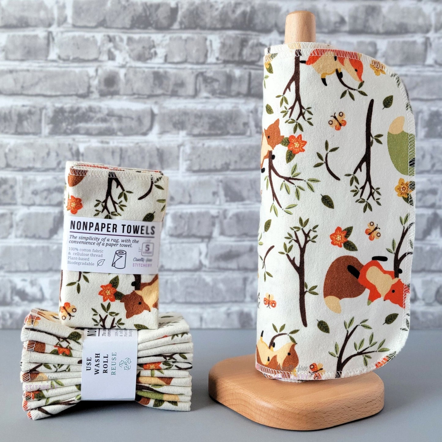 Ivory Foxes NonPaper Towels, displayed on a wooden paper towel holder next to packaged bundles. The print is an off-white or ivory background, with tree branches, orange flowers, and yellow and orange butterflies scattered around. There are a few foxes playing in the tree branches, some are orange, some are brown. 
