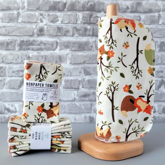 Ivory Foxes NonPaper Towels, displayed on a wooden paper towel holder next to packaged bundles. The print is an off-white or ivory background, with tree branches, orange flowers, and yellow and orange butterflies scattered around. There are a few foxes playing in the tree branches, some are orange, some are brown. 

