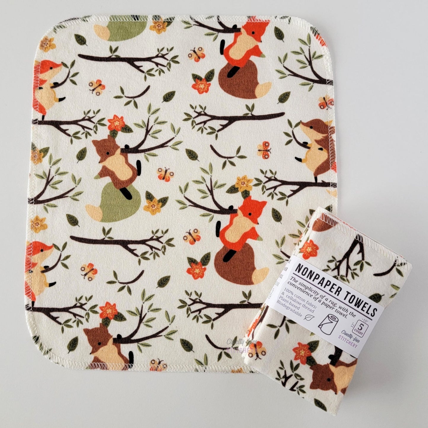 An unfolded Ivory Foxes NonPaper Towel, next to a bundled 5-pack. The towel is a rectangle with rounded corners, stitched around the edges with white thread. The bundled pack is folded up with a paper wrapper that says "NonPaper Towels, the simplicity of a rag with the convenience of a paper towel"
