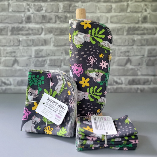 Koalas print NonPaper Towels. The print is gray koalas in various poses with their eucalyptus branches. Scattered around are simple pink and yellow flowers, all on a dark charcoal gray background. In the center, they are rolled onto a wooden paper towel holder, and there are bundled packs of 10 and 5 sitting next to the holder. The bundles are tied with string and have a small paper tag. The background is a gray brick wall and solid gray tabletop. 