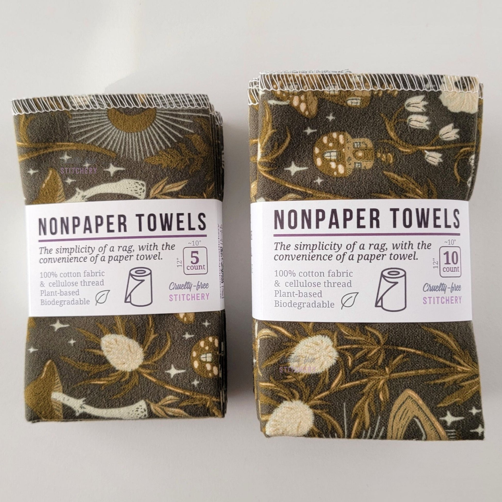 Two packages of the Lunar Forest NonPaper Towels, one 5-pack and one 10-pack. Both wrapped with a paper tag, The tag also has the fiber content, a graphic of a paper towel roll, the Cruelty-Free Stitchery logo, and a rectangle with rounded corners that has the dimensions on the outside, 10x12", and the number of towels inside.