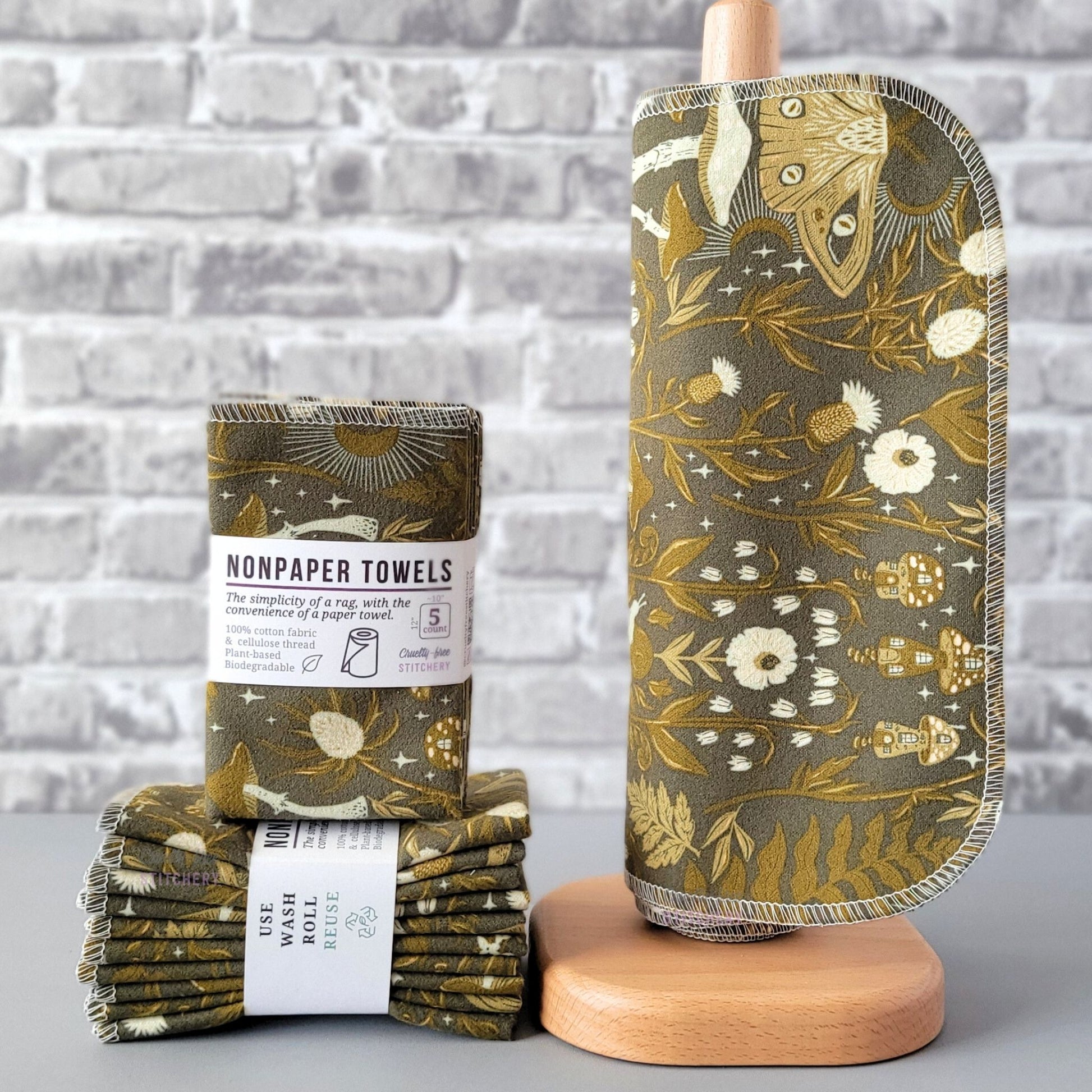 Lunar Forest NonPaper Towels, displayed on a wooden paper towel holder next to packaged bundles. The print is a very dark olive green with a variety of whimsical forest motifs all in lighter shades of olive green and off-white. Ferns, thistles, lots of mushrooms, some little mushroom houses, and a lunar moth. The print is large, so each separate NonPaper Towel may show different parts of the print.