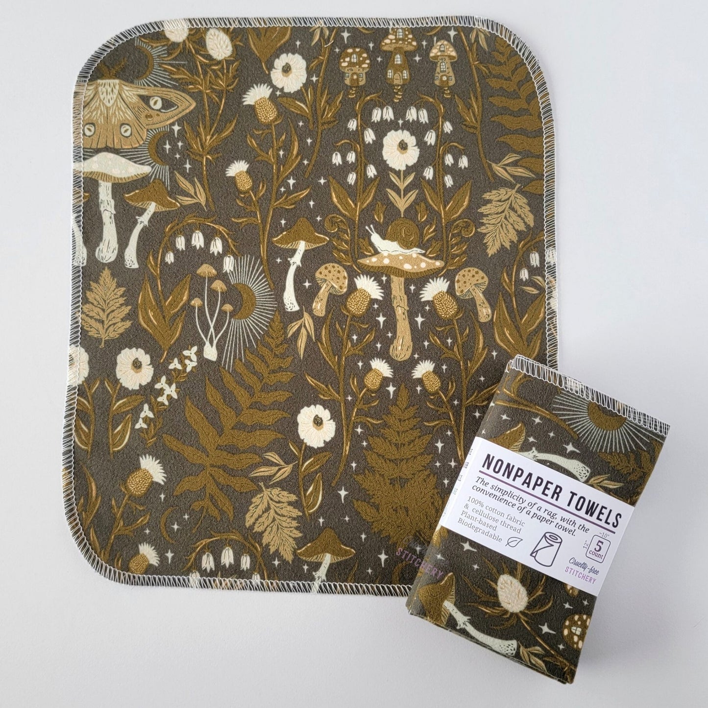 An unfolded Lunar Forest NonPaper Towel, next to a bundled 5-pack. The towel is a rectangle with rounded corners, stitched around the edges with white thread. This view of the print shows one of the mushrooms has a snail sitting on top. The bundled pack is folded up with a paper wrapper that says "NonPaper Towels, the simplicity of a rag with the convenience of a paper towel"
