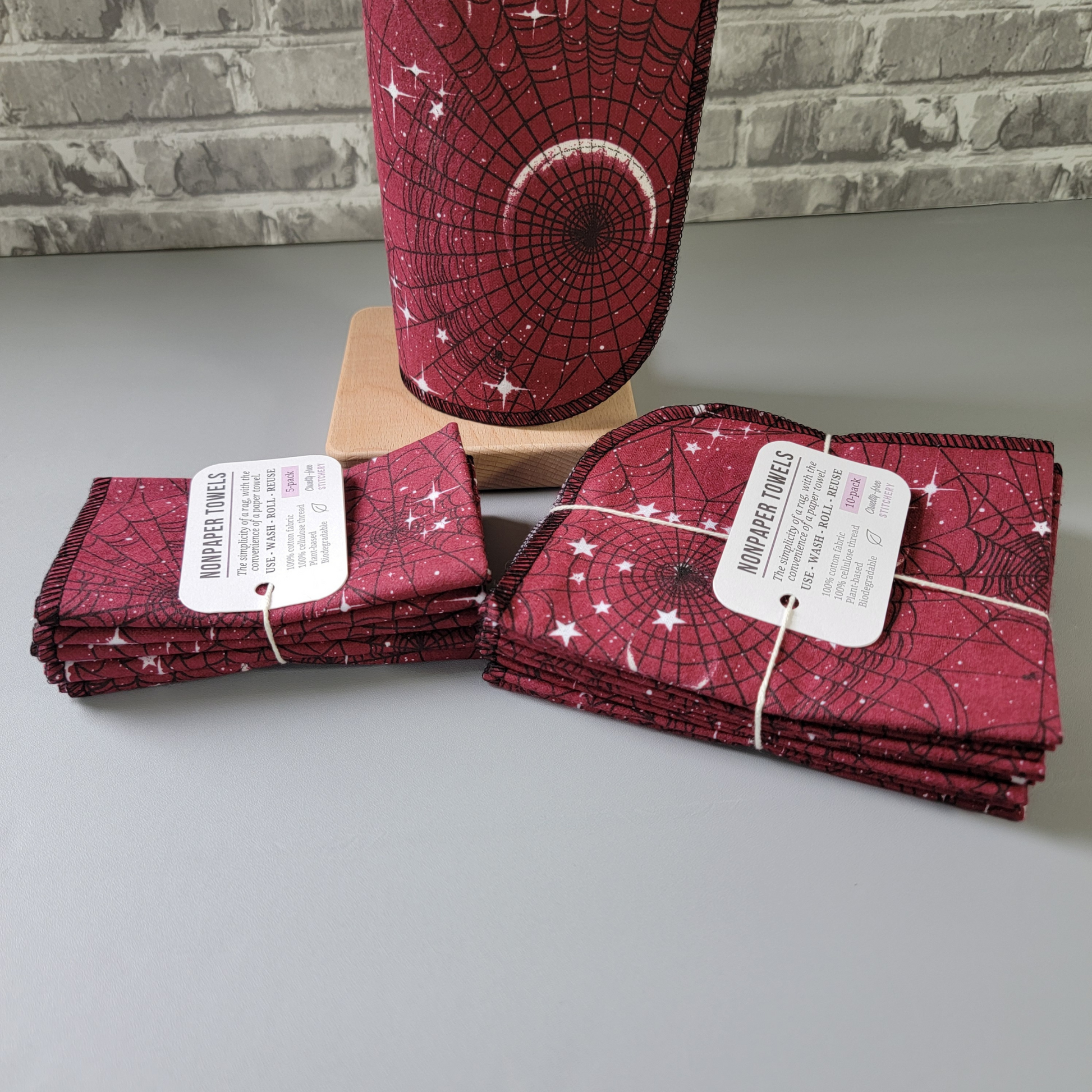 The red moon web nonpaper towels, a 5-pack and 10-pack laid side by side, photographed from the side to show the difference. The 5-pack is folded smaller than the 10-pack, both are tied with cotton string and a white paper tag. 