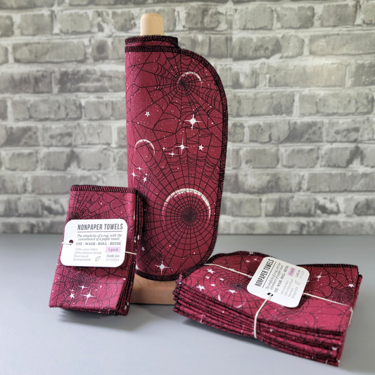 Red moon web NonPaper Towels, rolled onto a wooden paper towel holder, next to a 5-pack and 10-pack that are bundled with string and a tag. The towels are a deep cranberry red with black spider webs and white crescent moons and stars all over. 