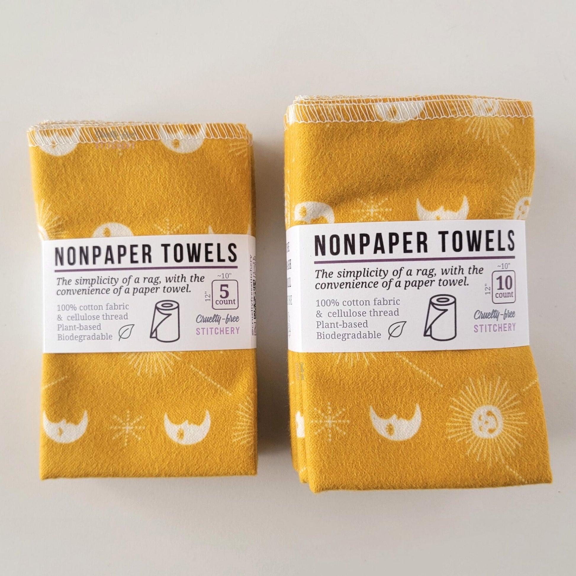 Two packages of the mustard celestial NonPaper Towels, one 5-pack and one 10-pack. Both wrapped with a paper tag, The tag also has the fiber content, a graphic of a paper towel roll, the Cruelty-Free Stitchery logo, and a rectangle with rounded corners that has the dimensions on the outside, 10x12", and the number of towels inside.