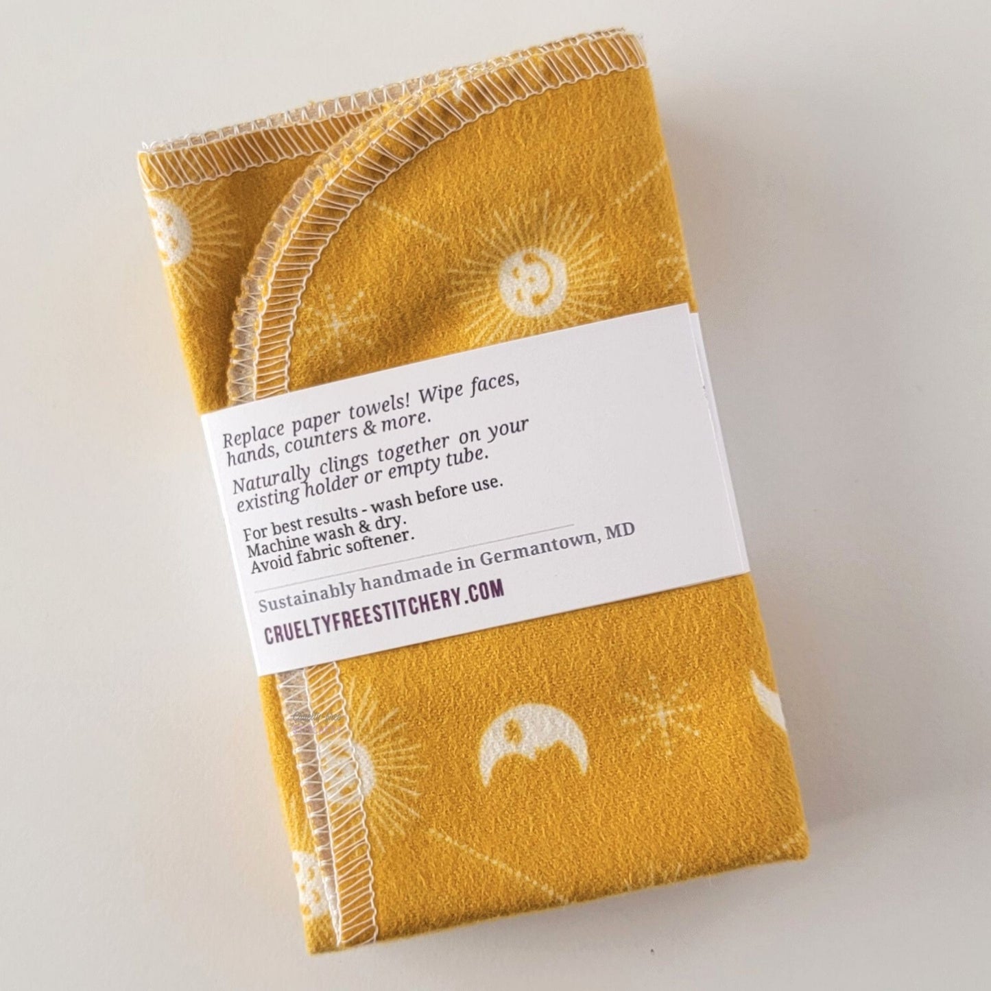The back of a packaged bundle of the yellow celestial NonPaper Towels. The tag reads "Replace paper towels! Wipe faces, hands, counters, and more. Naturally clings together on your existing holder or empty tube. For best results - wash before use. Machine wash and dry. Avoid fabric softener. Sustainable handmade in Germantown, Maryland. CrueltyFreeStitchery dot com."