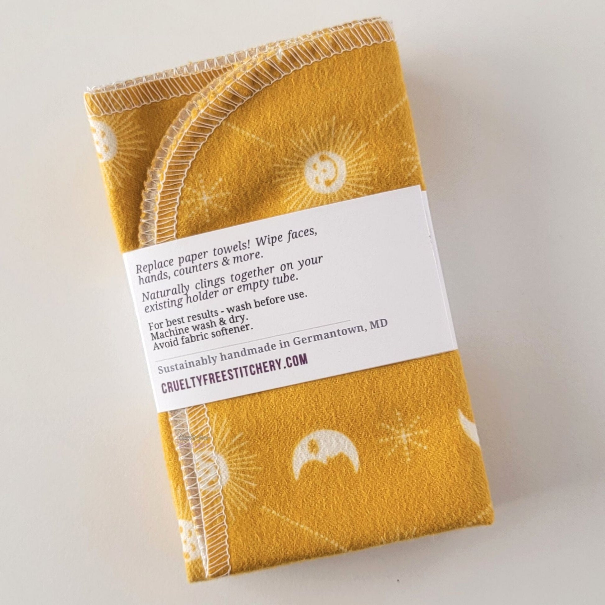 The back of a packaged bundle of the yellow celestial NonPaper Towels. The tag reads "Replace paper towels! Wipe faces, hands, counters, and more. Naturally clings together on your existing holder or empty tube. For best results - wash before use. Machine wash and dry. Avoid fabric softener. Sustainable handmade in Germantown, Maryland. CrueltyFreeStitchery dot com."