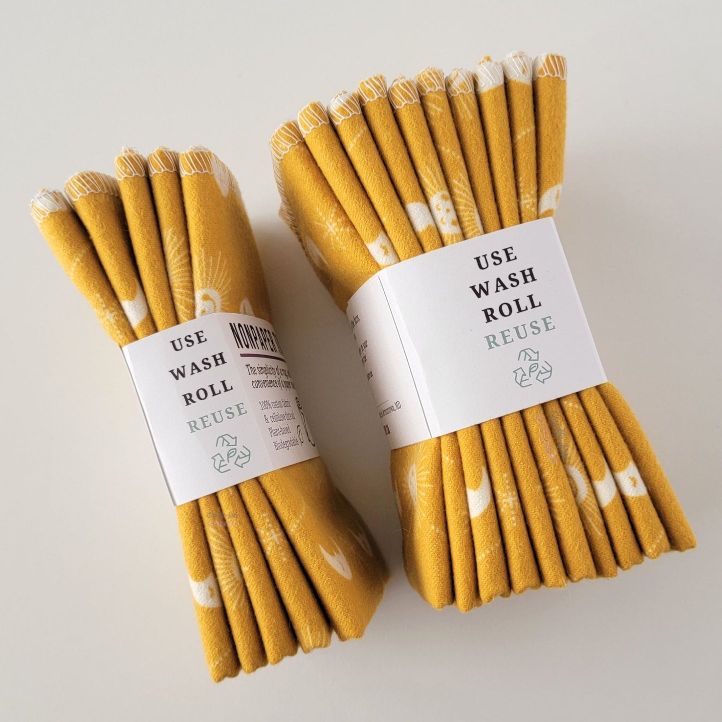 Two packages of the mustard celestial NonPaper Towels, shown on their side to display the 5-pack and 10-packs. The side of the paper tag says "use, wash, roll, reuse" and has the recycling logo with a leaf in the center, to symbolize they are biodegradable.