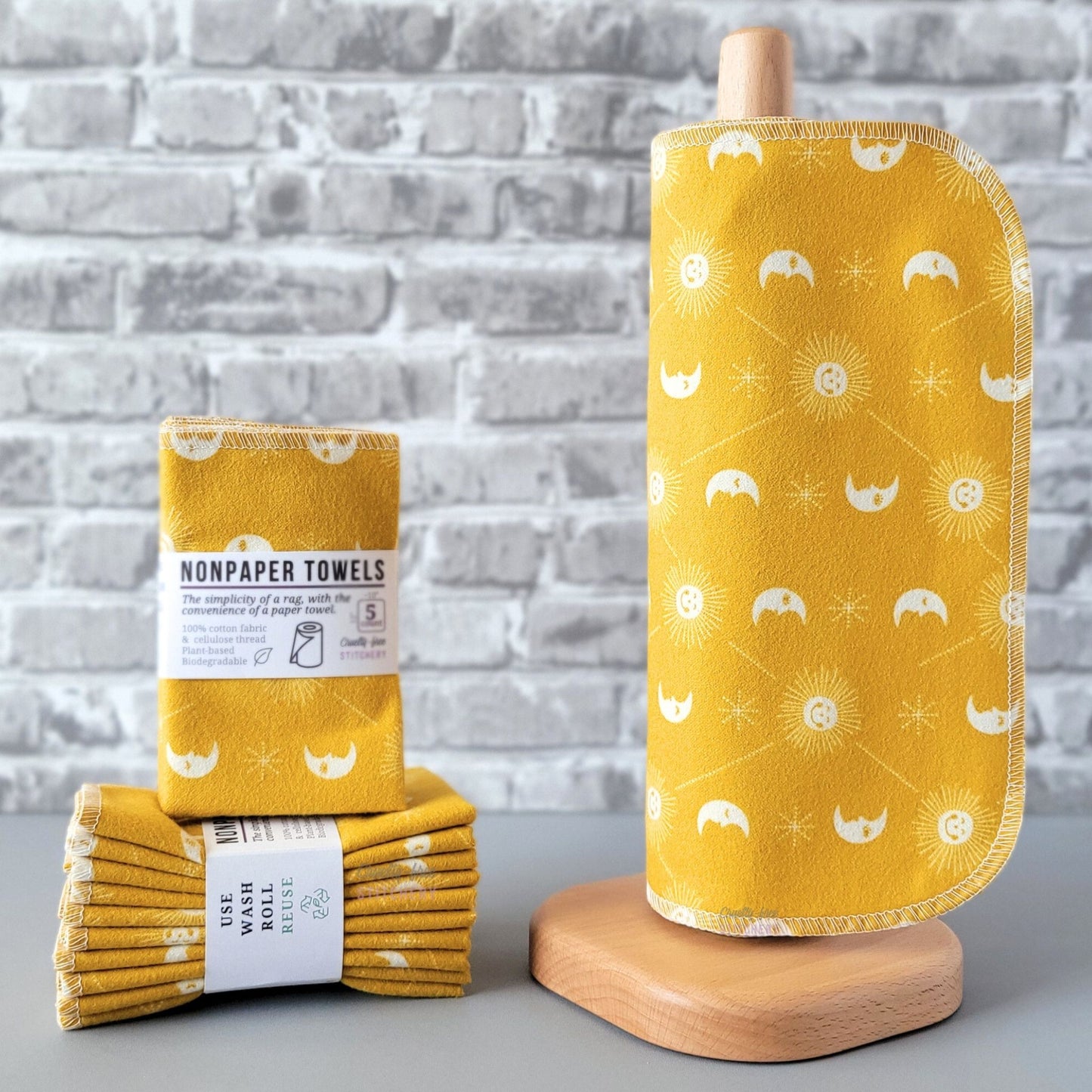 Mustard yellow celestial NonPaper Towels, displayed on a wooden paper towel holder next to packaged bundles. The print is a bold mustard yellow background, with small smiling sunshines, sleepy crescent moons, and tiny dotted star shapes, with thin diagonal lines between the suns. All printed in white.