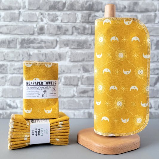 Mustard yellow celestial NonPaper Towels, displayed on a wooden paper towel holder next to packaged bundles. The print is a bold mustard yellow background, with small smiling sunshines, sleepy crescent moons, and tiny dotted star shapes, with thin diagonal lines between the suns. All printed in white.